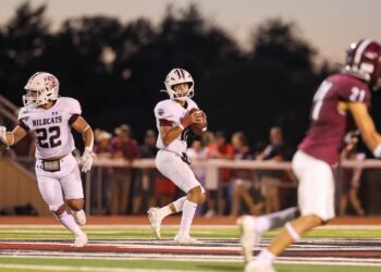 Seabolt Chase-14651 | Press Pass Sports Quarterback in white Wildcats uniform with ball, looking for a path as a teammate runs behind him on a football field at dusk, a maroon defender in foreground left.