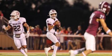 Quarterback in white Wildcats uniform with ball, looking for a path as a teammate runs behind him on a football field at dusk, a maroon defender in foreground left.