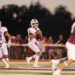 Seabolt Chase-14651 | Press Pass Sports Quarterback in white Wildcats uniform with ball, looking for a path as a teammate runs behind him on a football field at dusk, a maroon defender in foreground left.