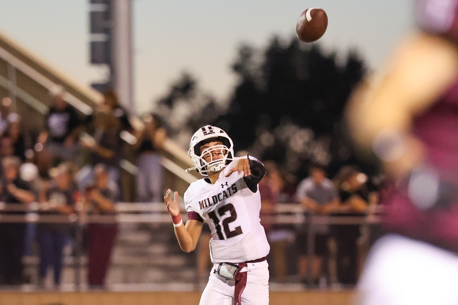 Football quarterback in a white Wildcats jersey number 12 throws a pass on a high school field with cheering crowd in the stands behind him