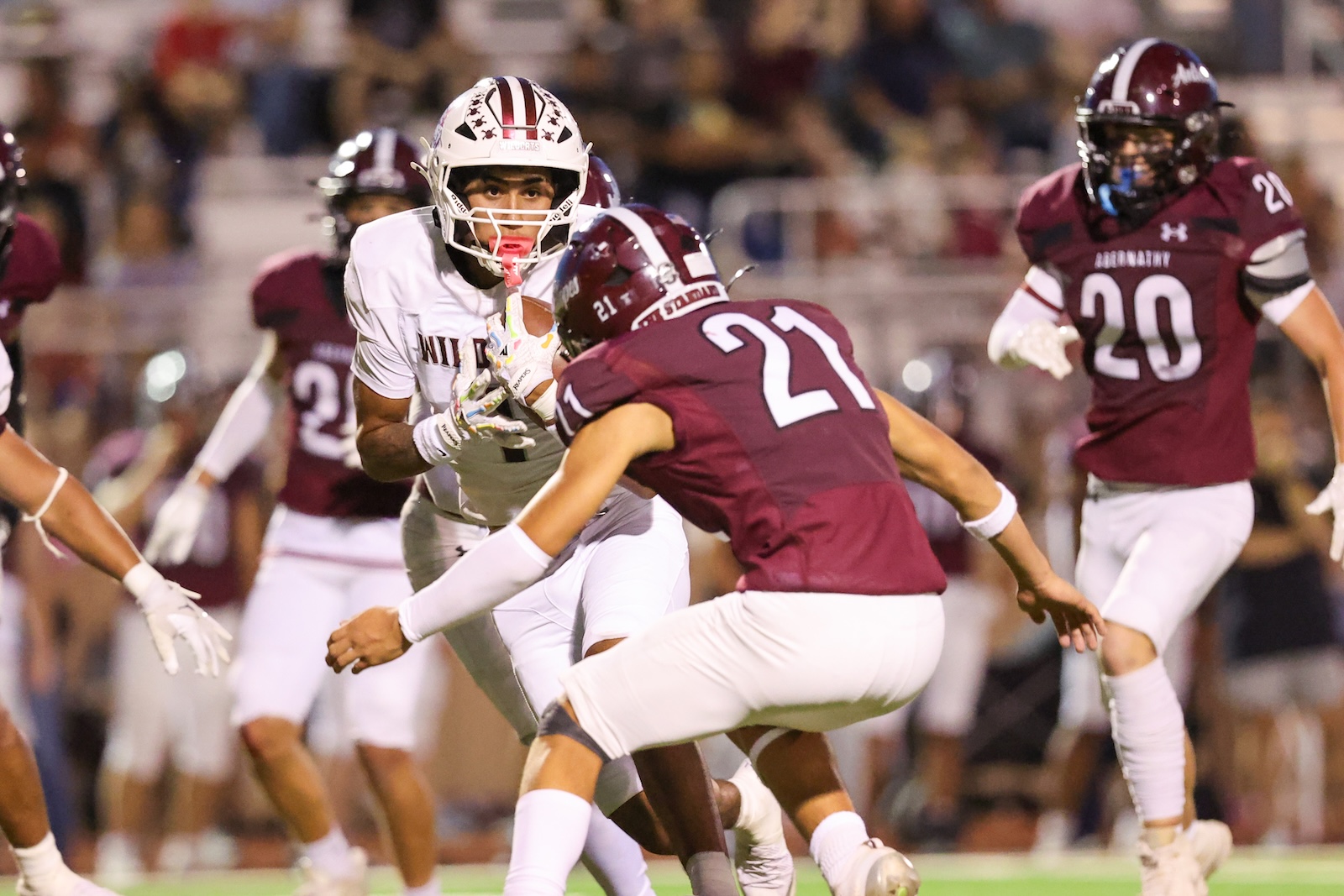 Football game action: player in white with the ball faces a tackler in maroon (#21) during a nighttime play with other players in maroon nearby.