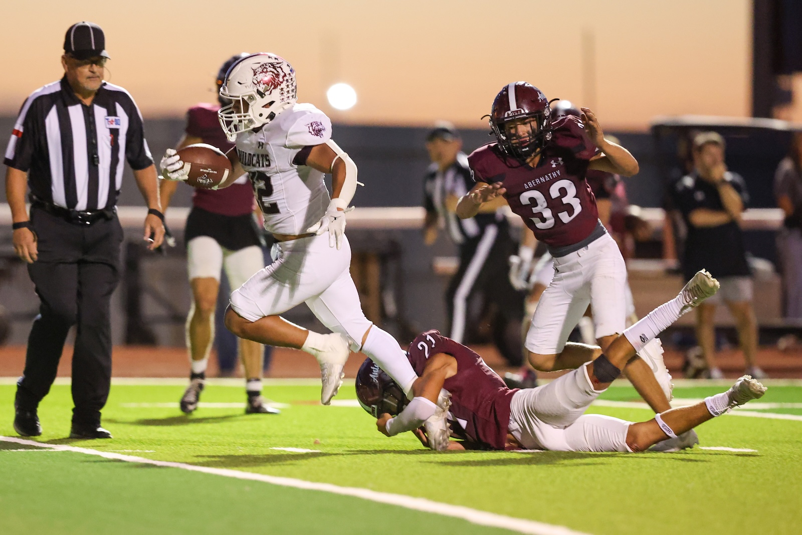 Football player in a white Wildcats uniform runs with the ball as a maroon-clad defender dives to tackle him behind.