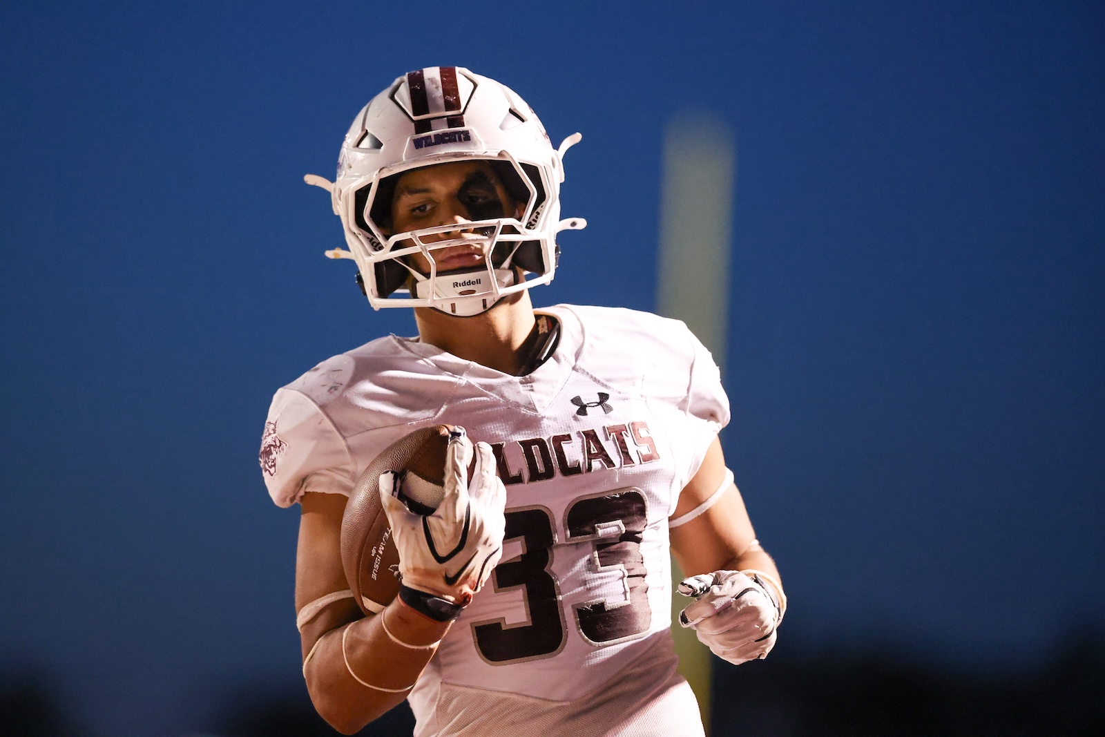Football player in a white Wildcats jersey and helmet running with the ball, gripping it with one hand and looking ahead along the field.