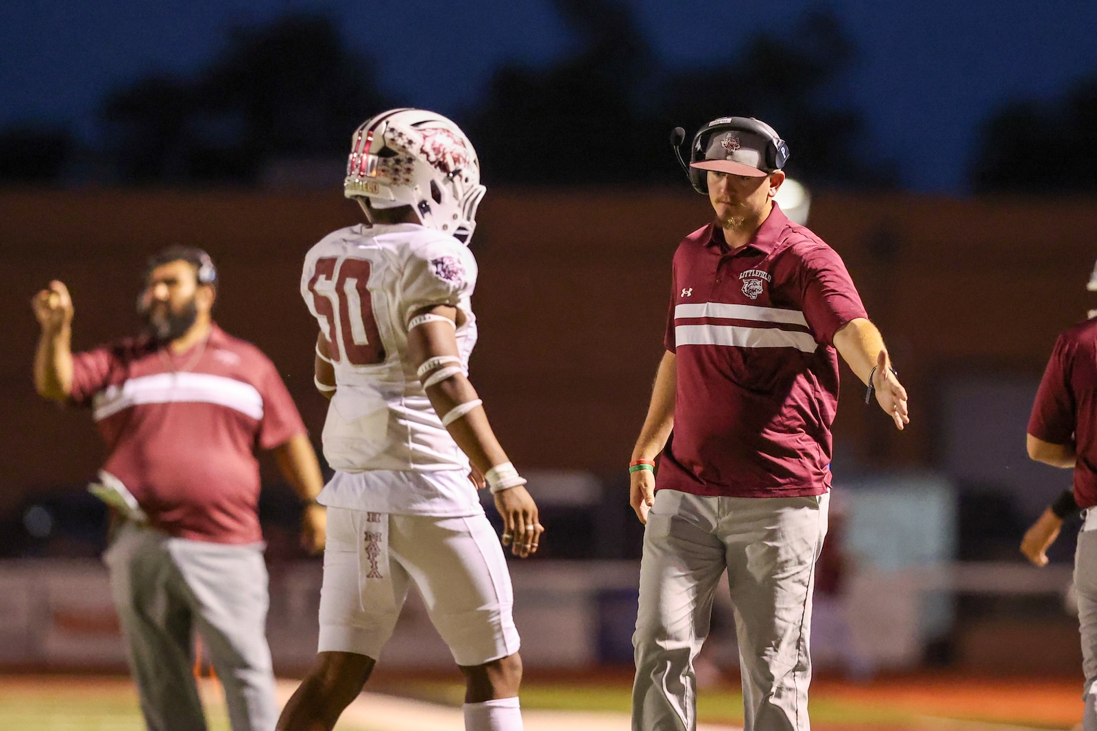 Football coach in a maroon polo with headset extends a hand toward a white-jersey player on a night practice or game field.