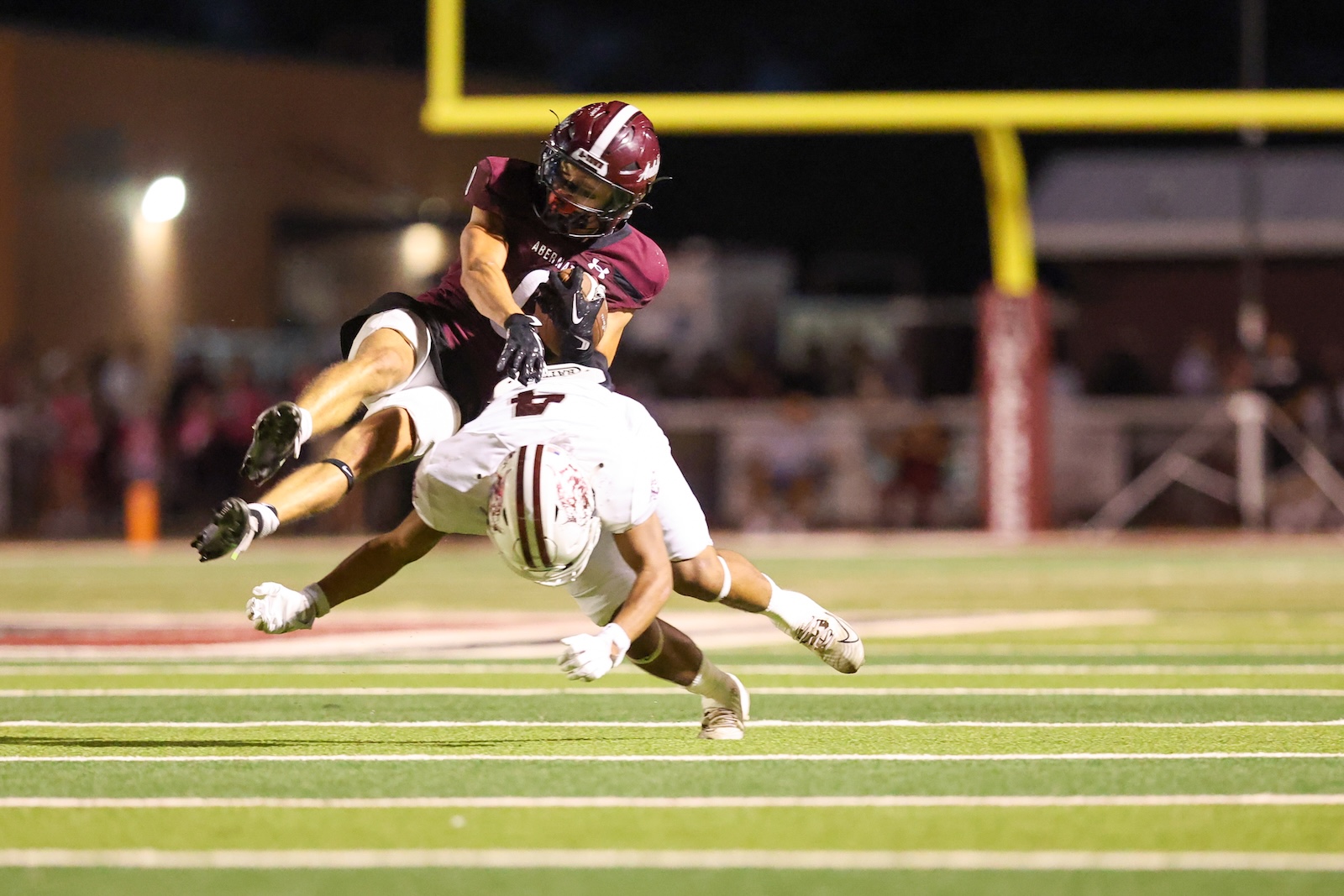 Two football players collide on a night game, maroon jersey tackling a white-shirted opponent near the end zone.