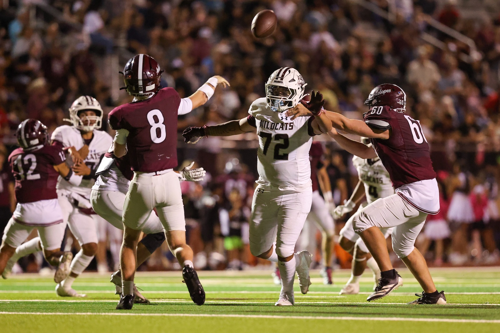 Night game action: a Wildcats player in white (#72) extends to catch a high pass while maroon-clad defenders converge nearby with the ball in midair above them.