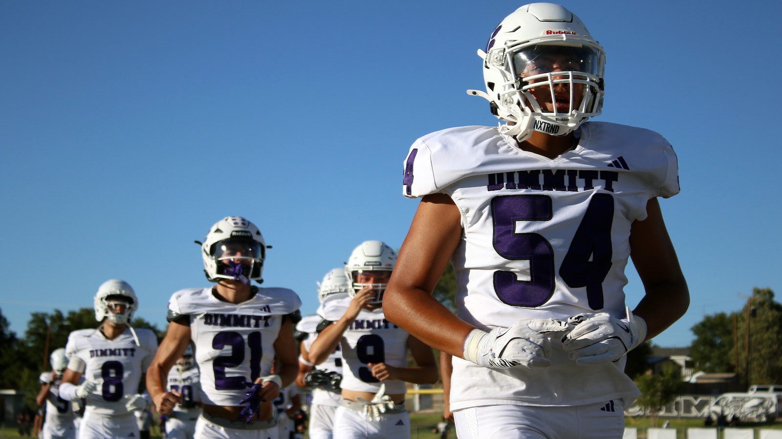 Young football players in white uniforms with purple numbers sprint onto the field, Dimmitt team name visible on jerseys under a clear blue sky.