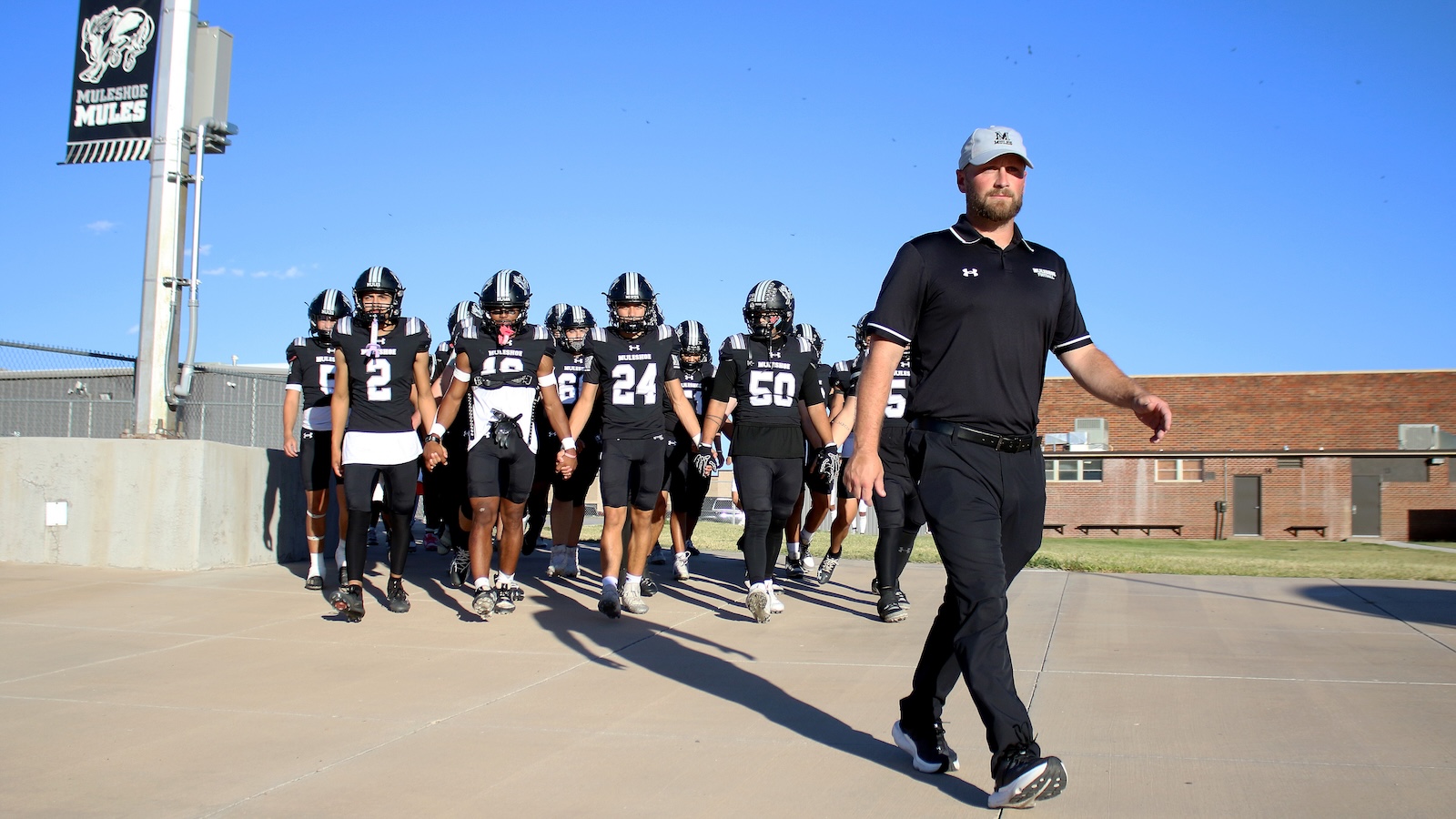 Football coach leads a black-uniformed high school team onto the field, players marching behind him under a clear blue sky.