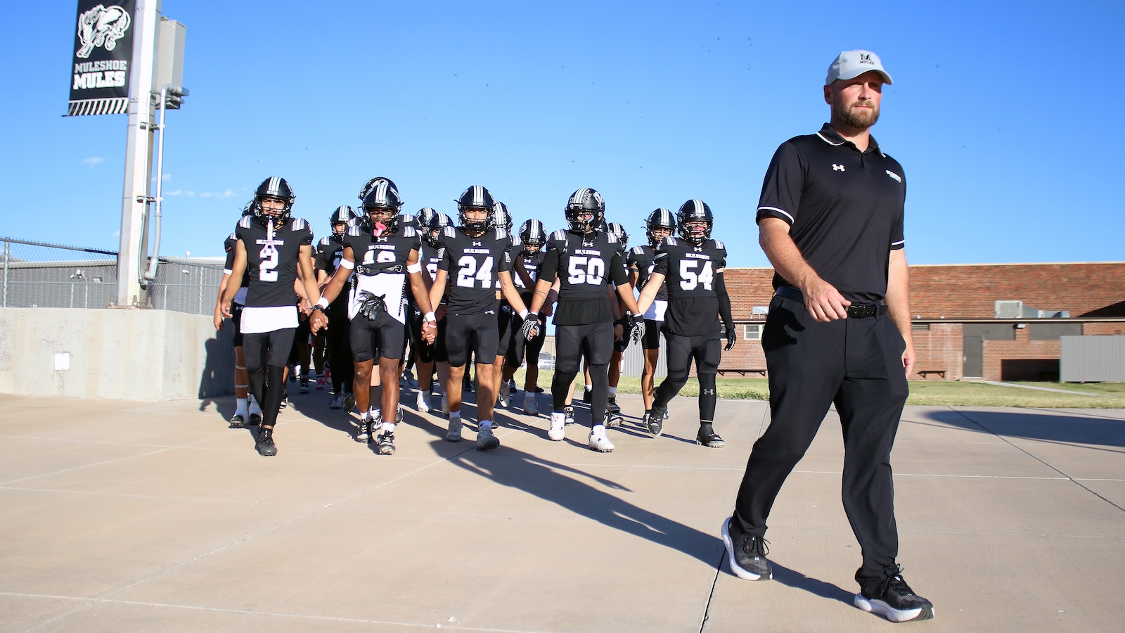 Black-clad football players walk onto the field behind a coach in a black polo shirt and gray cap on a sunny day with a banner in the background.
