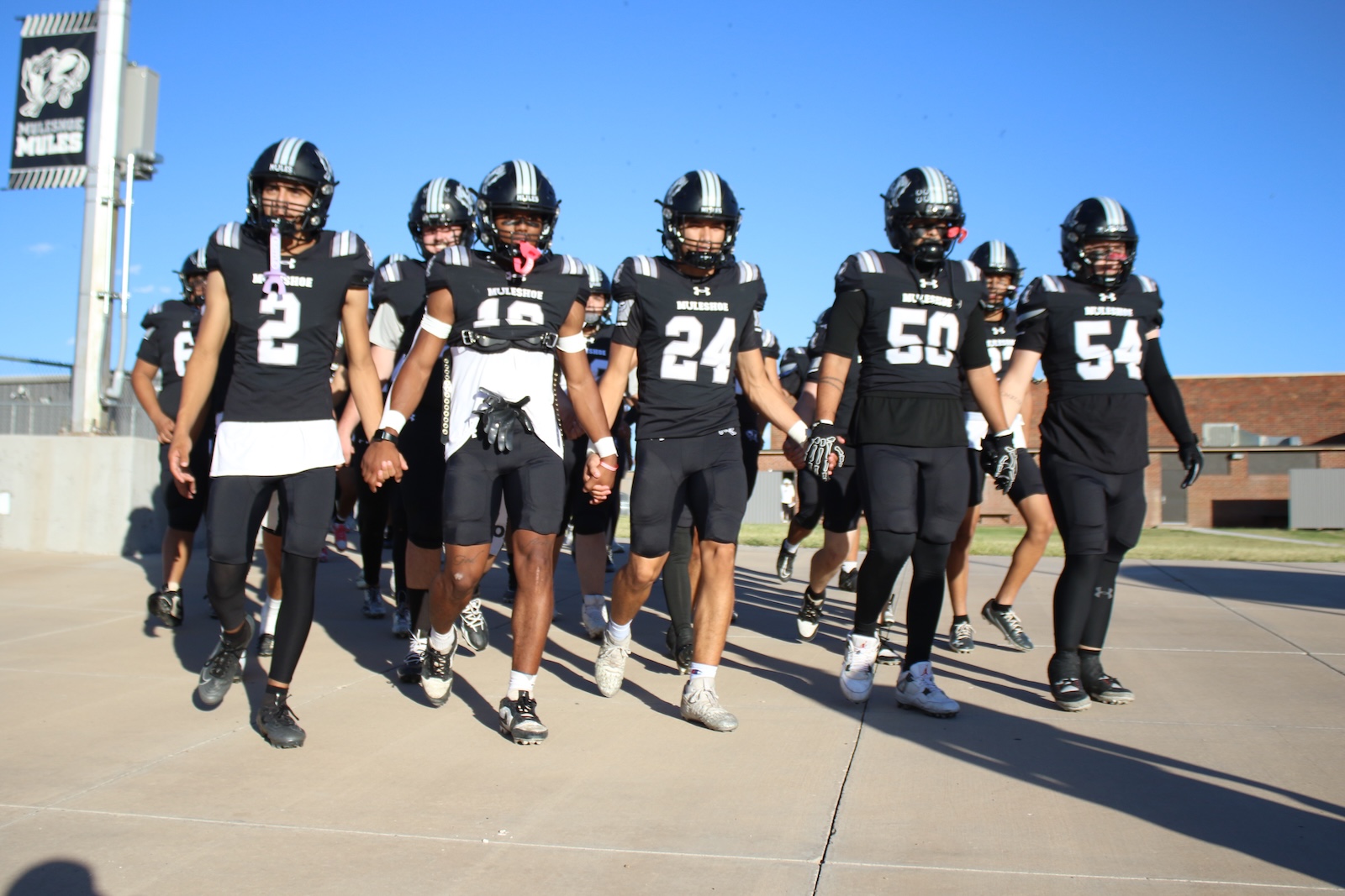 High school football team in black uniforms walk together hand in hand toward the camera on a sunny day.