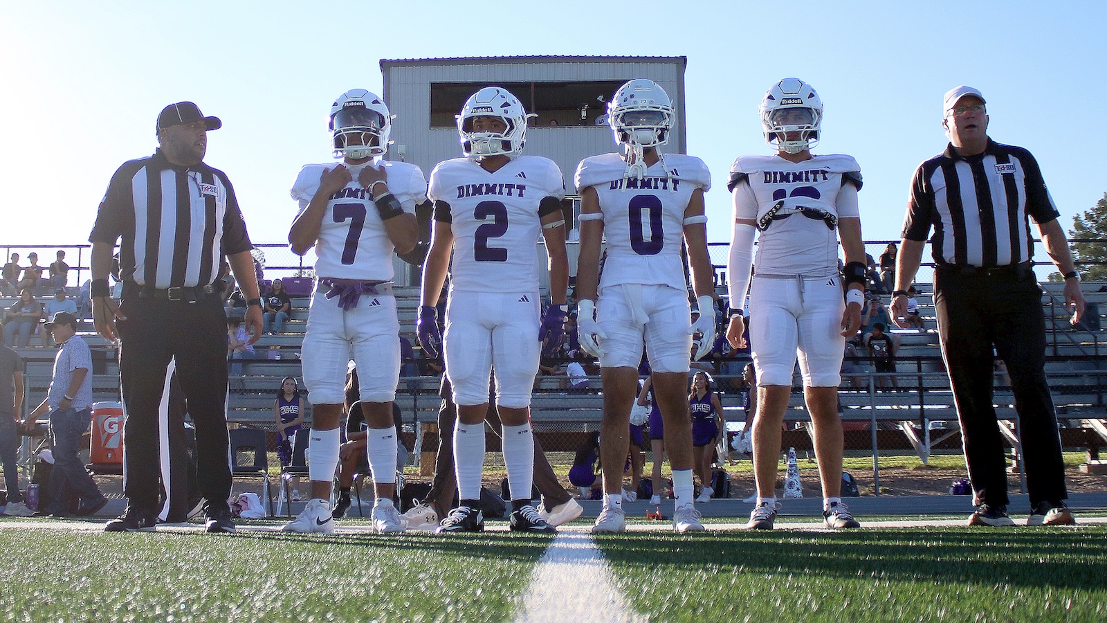 Five football players in white uniforms and helmets stand in a line on the field, numbers 7, 2, and 0 visible, flanked by referees in black-and-white stripes.