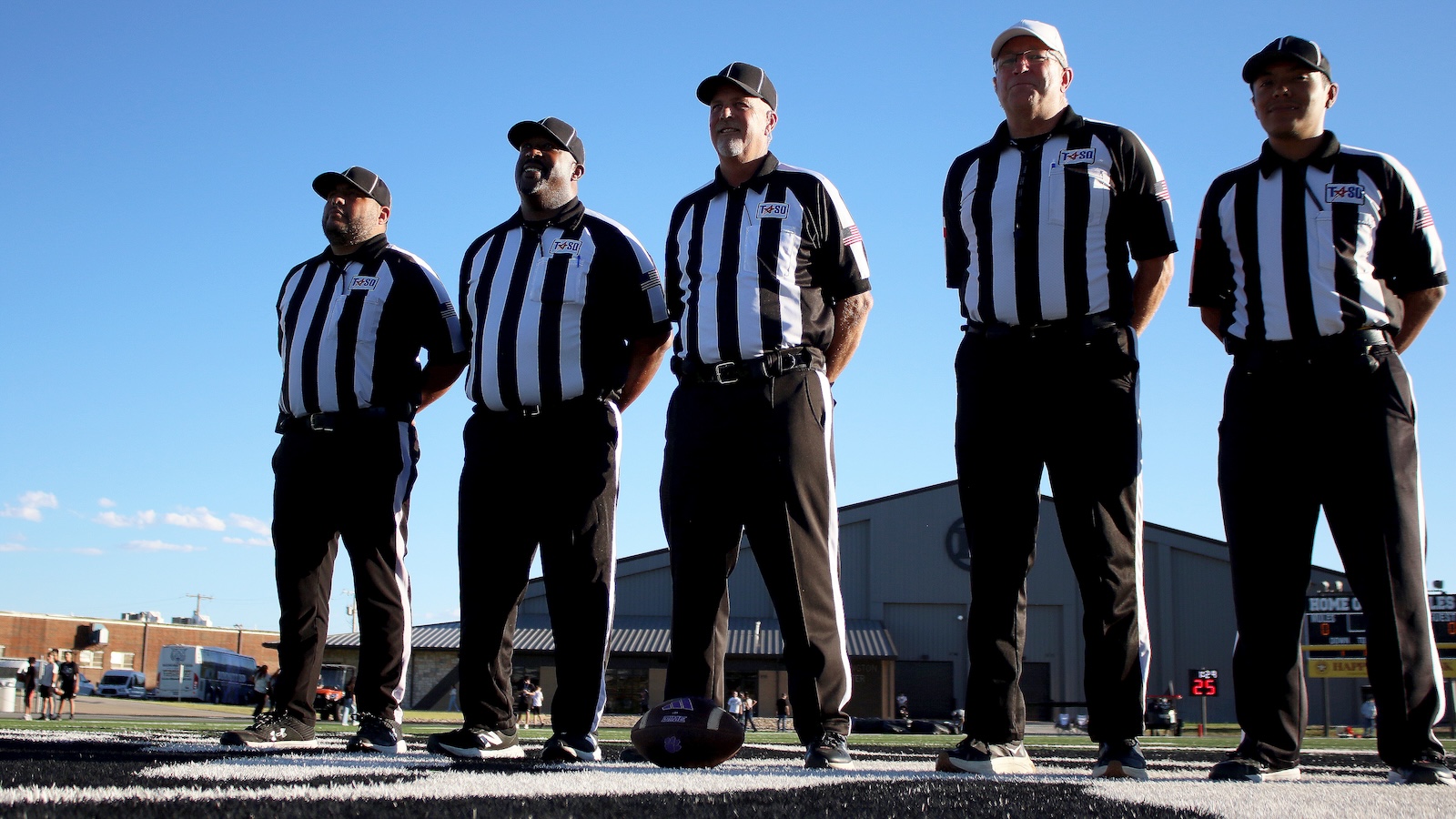 Five referees in black-and-white striped uniforms stand in a line on the field, with a football at their feet against a clear blue sky.