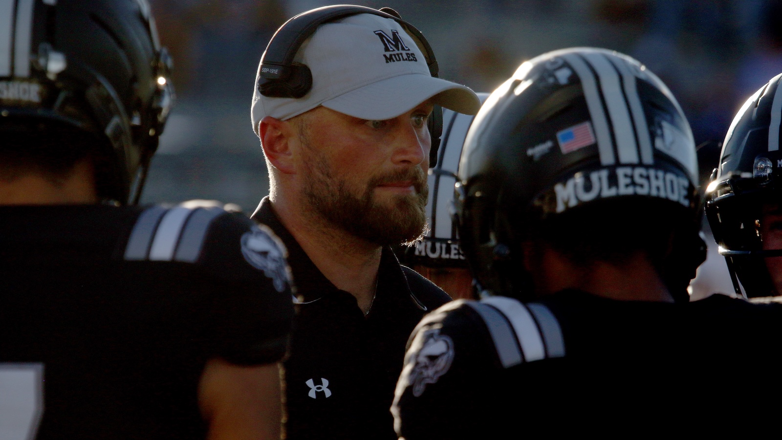 Football coach with a white cap and headset talking to his team on the sideline.