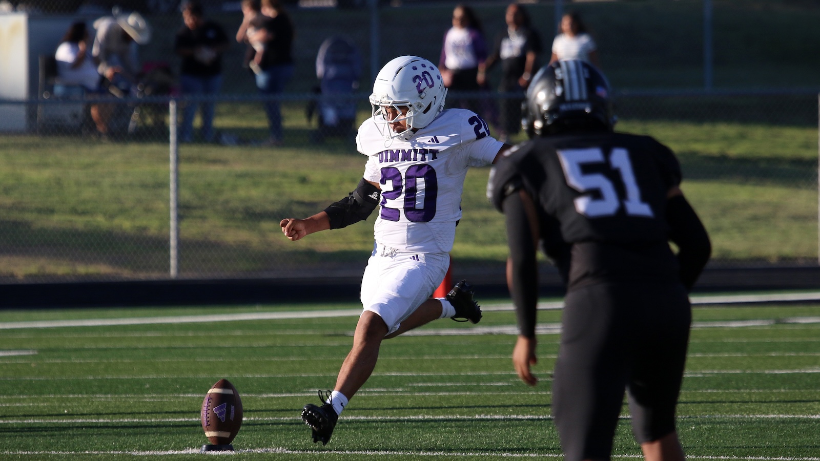 Football player in white jersey #20 kicking the ball on a green field as a defender in black #51 closes in.