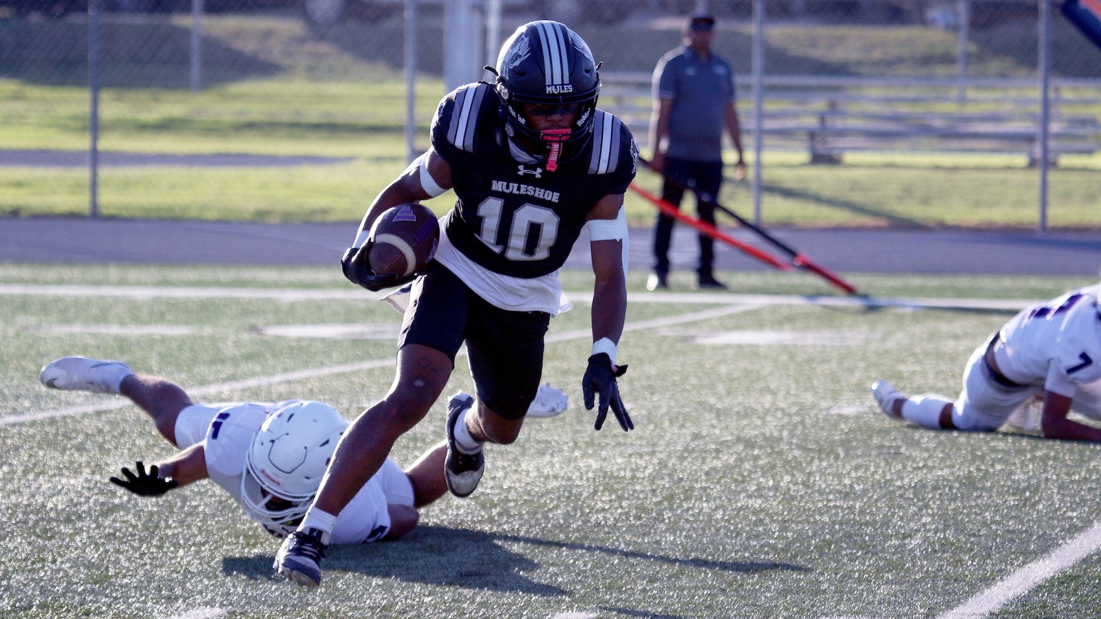 Football player in a black jersey (#10) sprints with the ball, dodging white-uniform tacklers on a turf field.