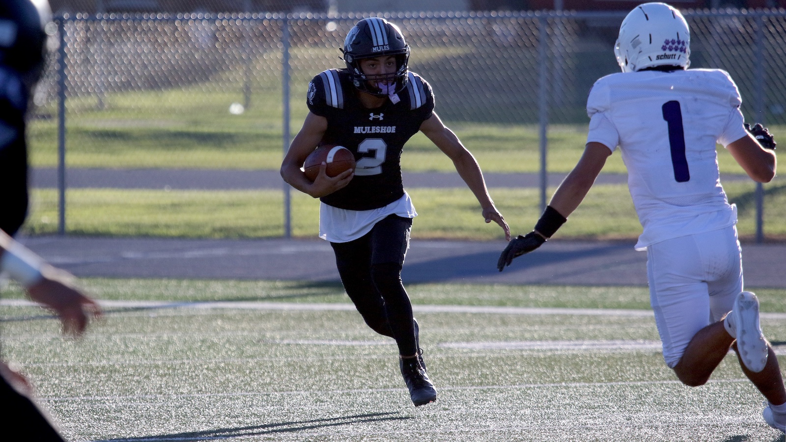 Football player in a black Muleshoe jersey (number 3) runs with the ball, chased by a defender in white on a sunny field.