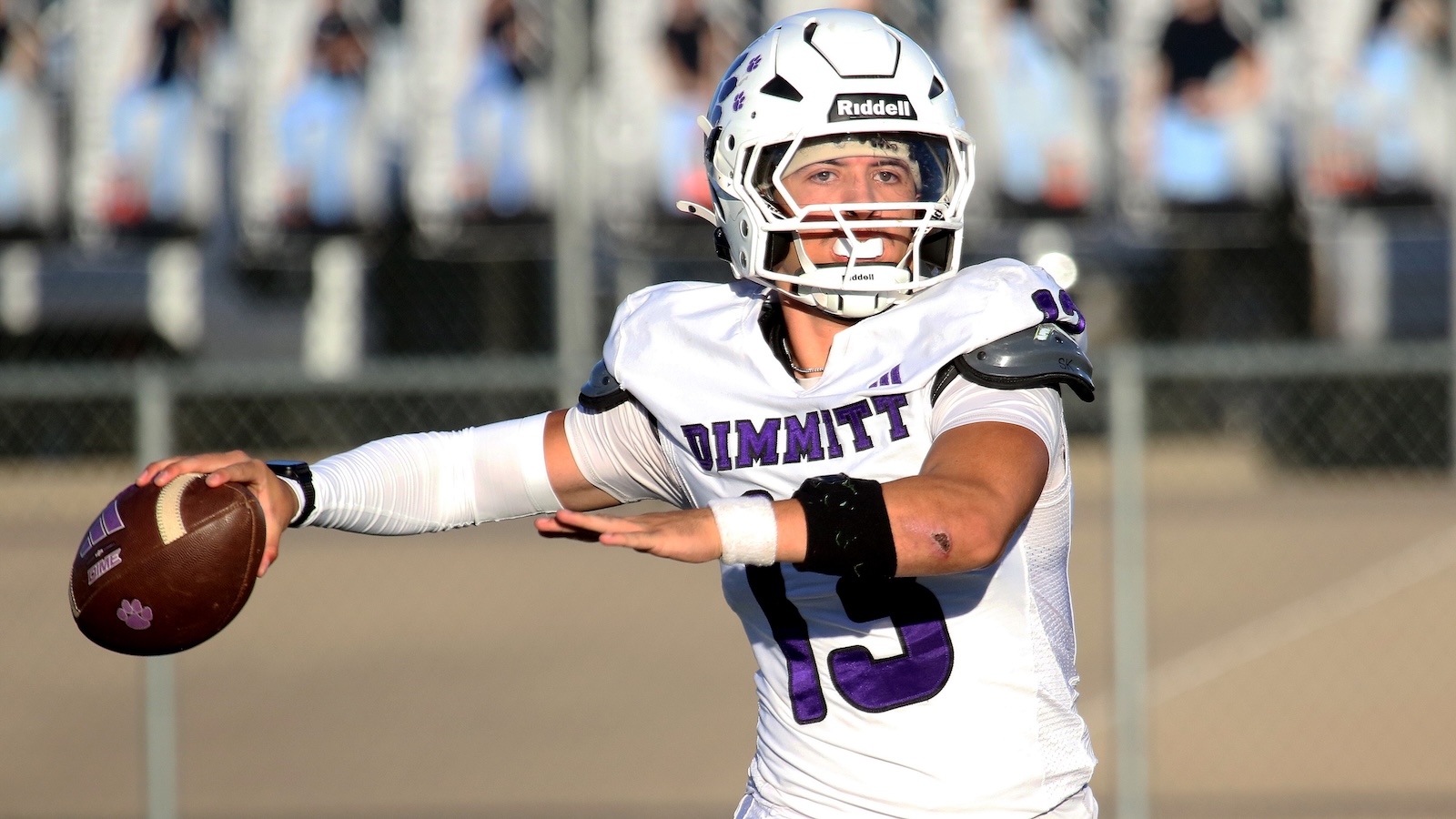 Quarterback in a white Dimmitt jersey (#15) throwing a football during a game, helmet on, on a sunny field with spectators in the background.