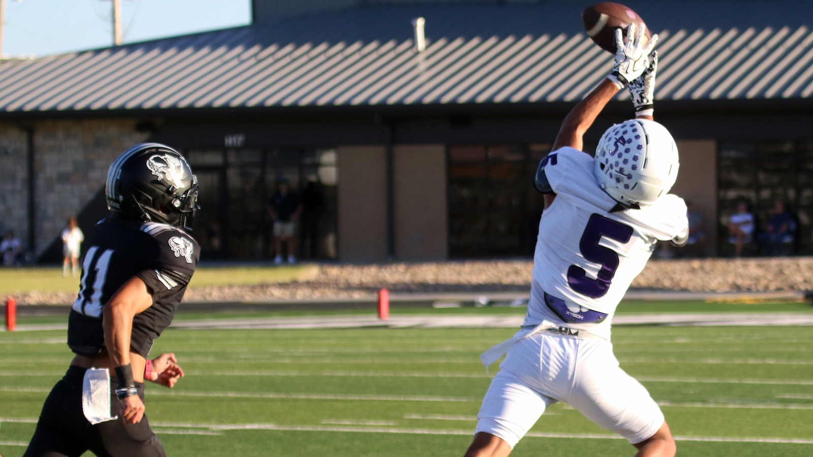White-clad football player, number 5, leaps to catch a football as a defender in black watches on a sunny field.