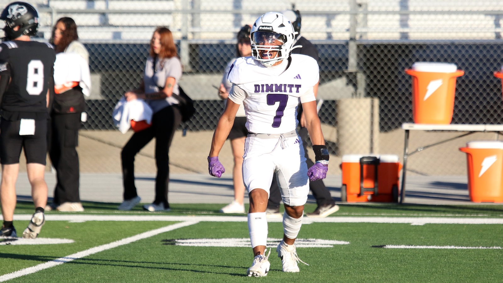 Football player in a white uniform (#7) on a green turf, wearing a helmet and purple gloves, standing on the field with teammates nearby.