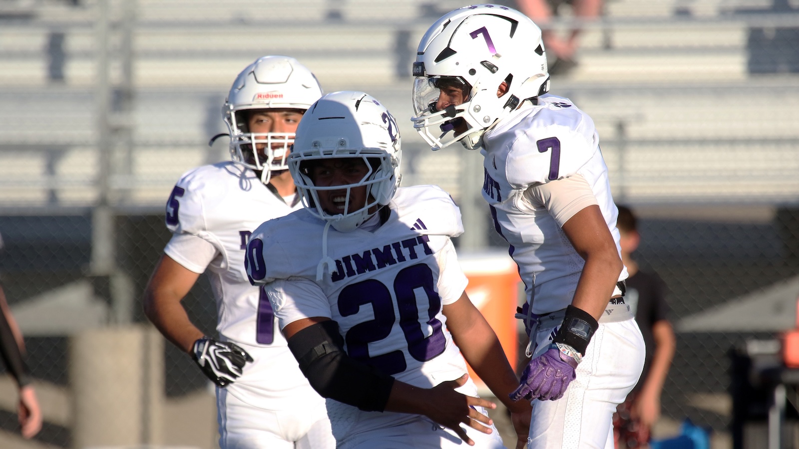 Three football players in white and purple uniforms celebrate on the field during a game.