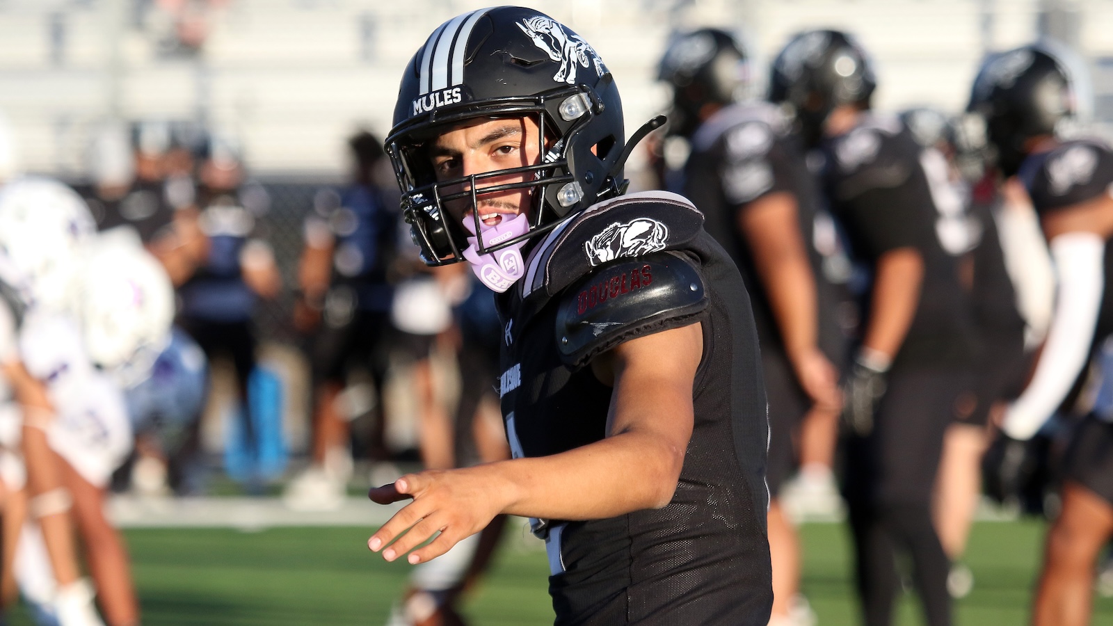 Football player in a black uniform extends an arm on a football field during a game, teammates in the background.