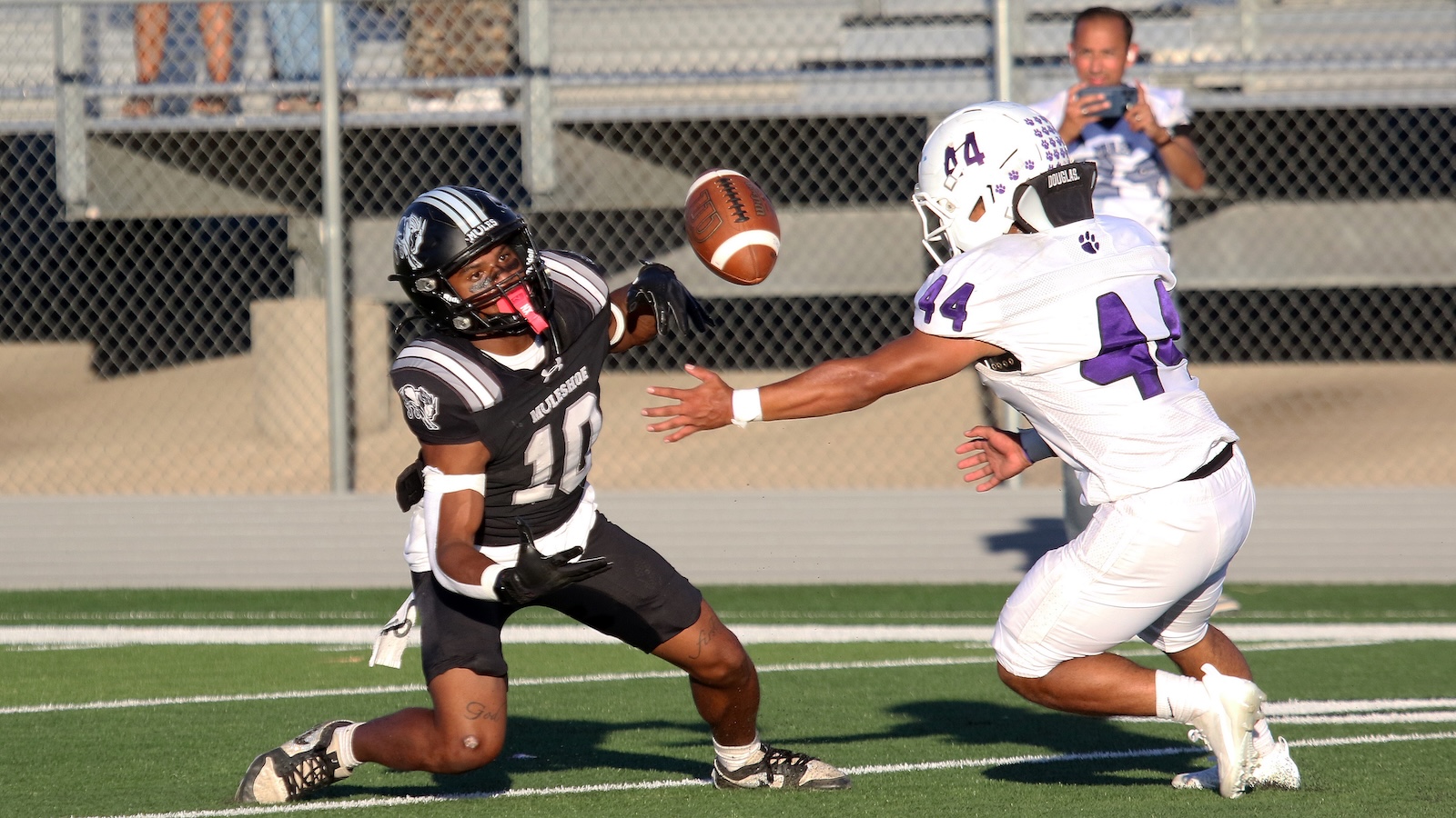 Two football players in black and white jerseys reach for a spiraling ball on a sunny field; a chain-link fence and bleachers in the background.