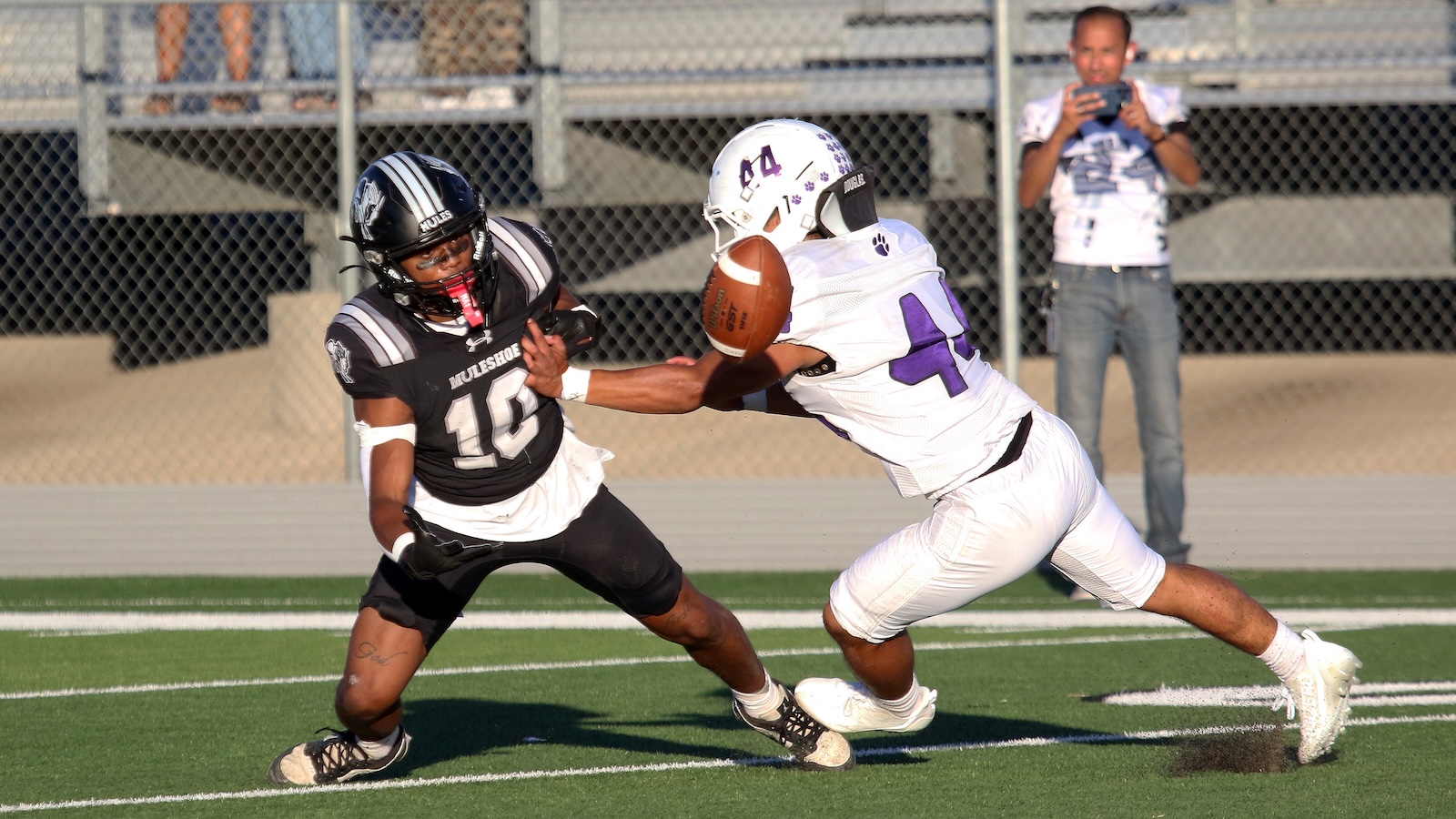 Two football players collide for the ball on a green turf field, black jersey #10 on the left and white jersey #4 reaching for the ball while a spectator films in the background.