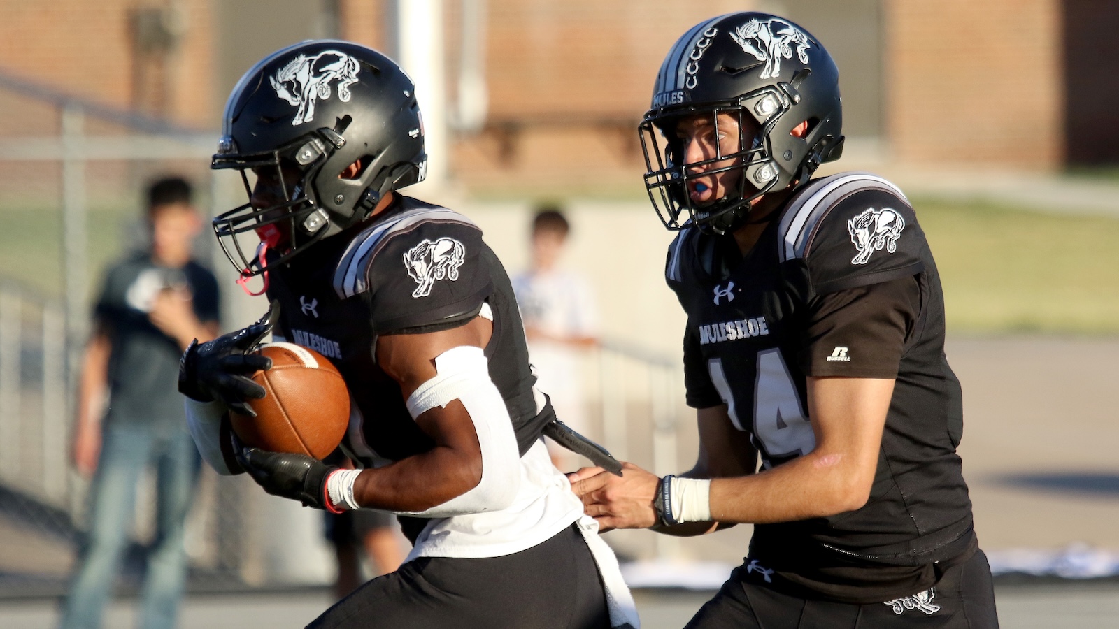 Two football players in black uniforms collide on the field, one carrying the ball as a teammate reaches in to help or defend him.