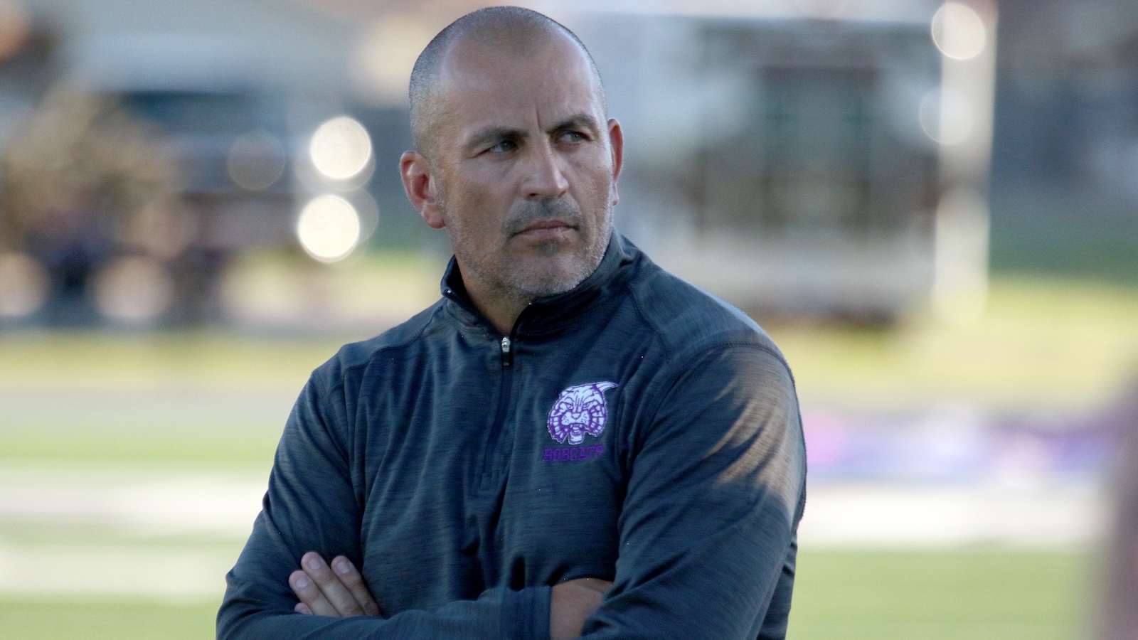 The man with a shaved head and stubble, wearing a dark zip-up jacket with a purple dragon logo, stands outdoors with arms crossed and a serious expression.