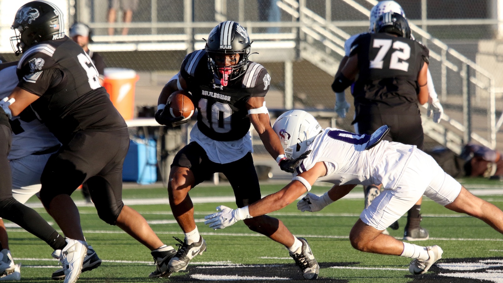 Football player in black uniform runs with the ball while a defender in white stretches out to tackle him on a green field with bleachers in the background