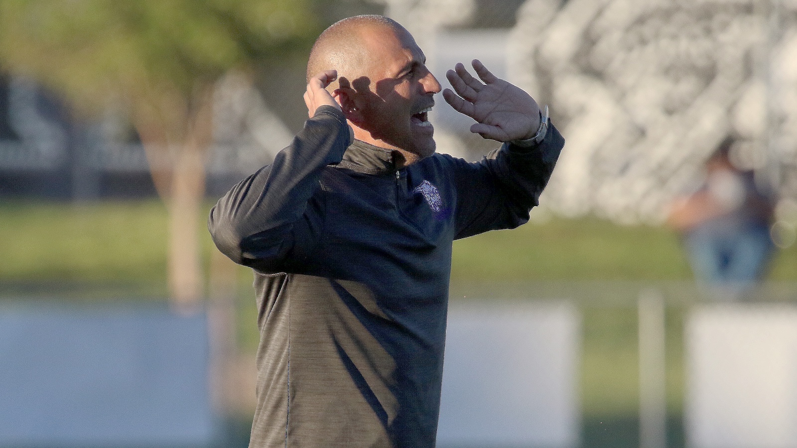 Man in a dark athletic shirt shouts with his hands cupped around his mouth on a sports field.