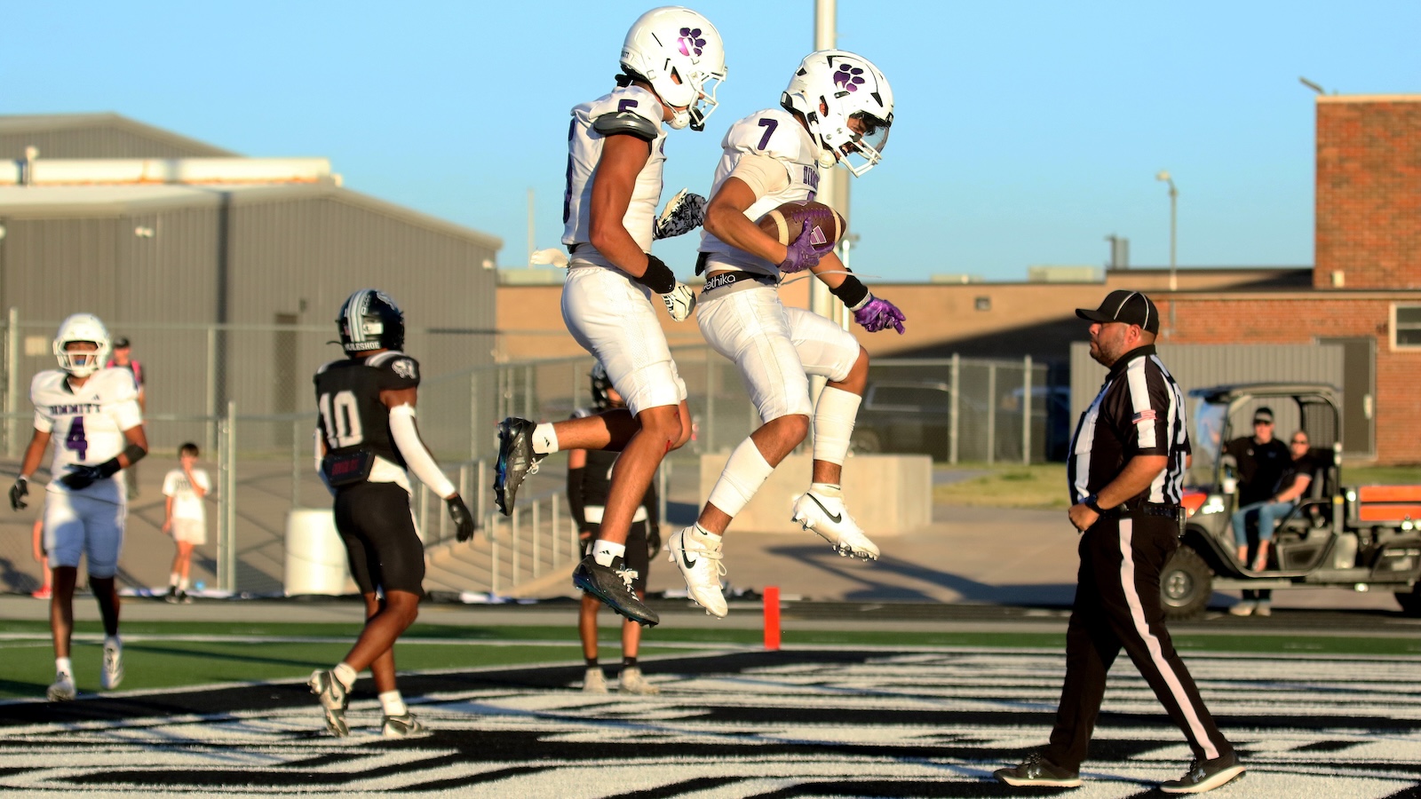 Two football players in white uniforms leap mid-air as one holds a ball, celebrating near the end zone while a referee watches.