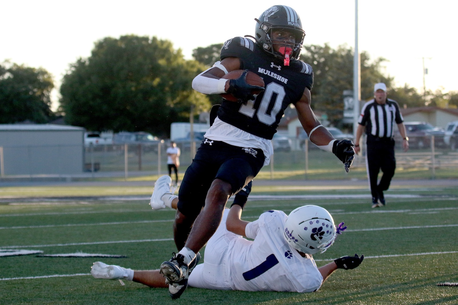 Black-clad football player (number 10) charges forward with the ball as a defender falls at his feet on a turf field, a referee in the background.