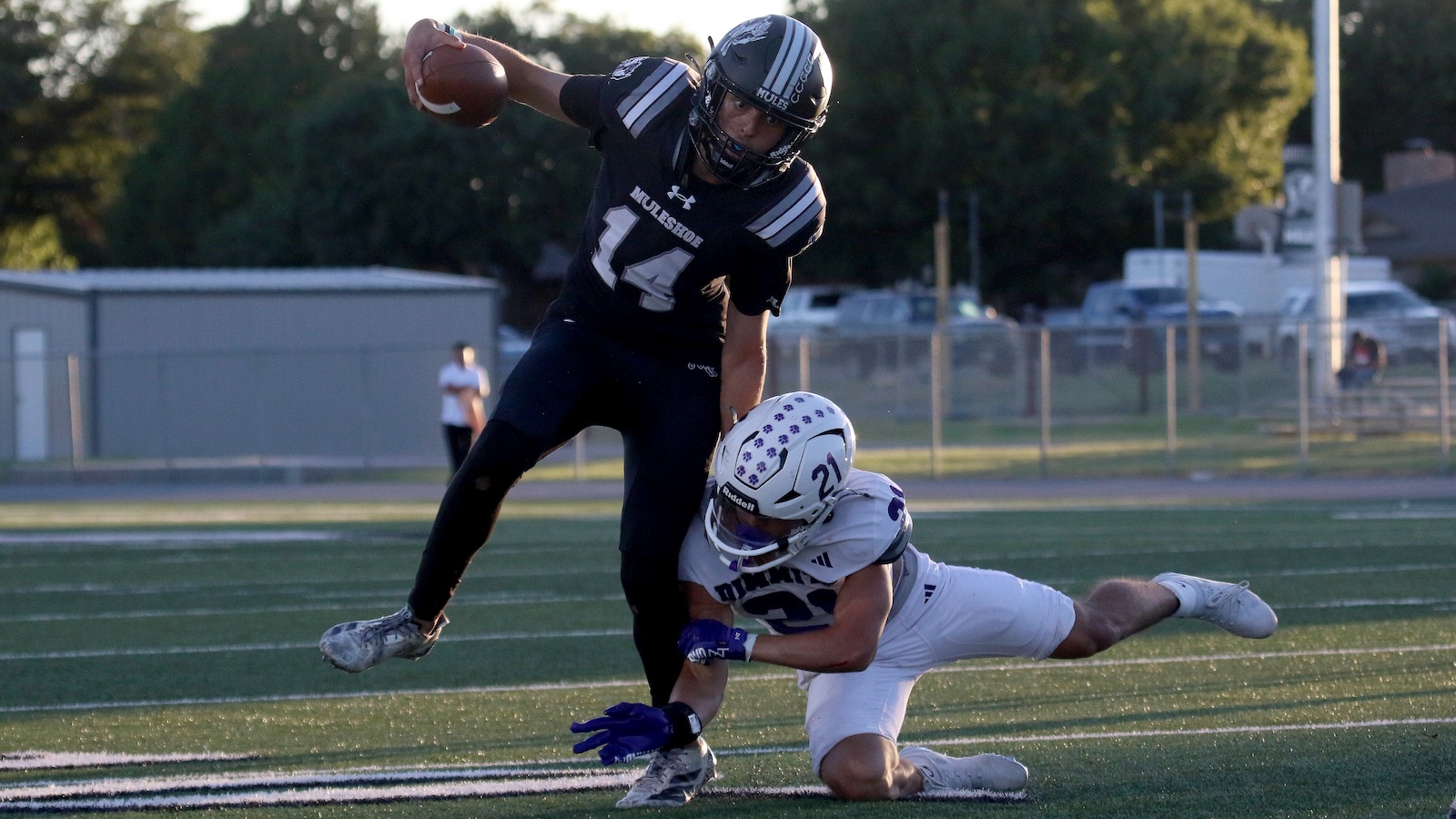 Football action: a player in a black uniform leaps with the ball as a defender in white dives to tackle nearby on a turf field.