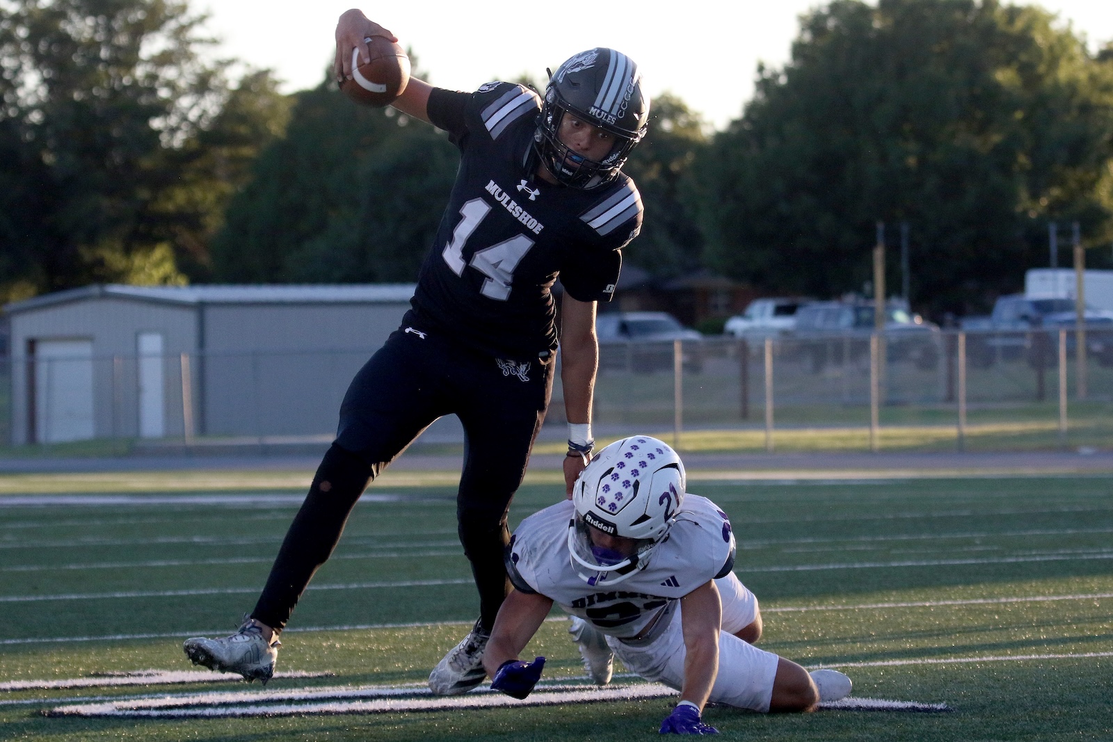 Football player in black jersey (#14) lunges to throw a pass while a white-uniformed opponent falls on the field behind him.