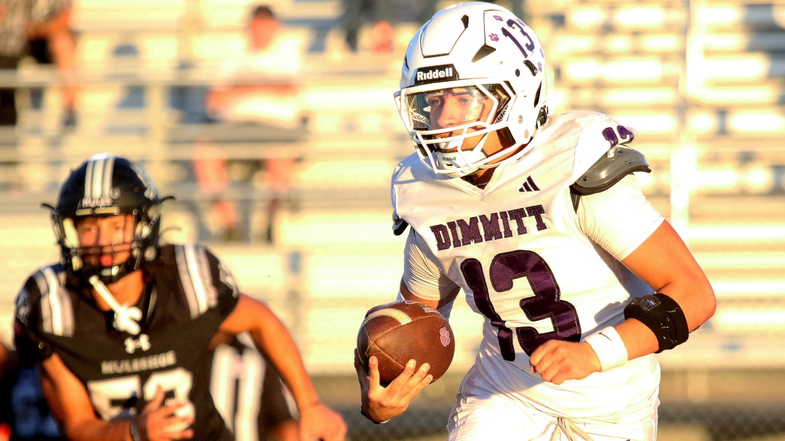 American football player wearing a white Dimmit #13 jersey carries the ball downfield as a black-clad defender pursues in the background.