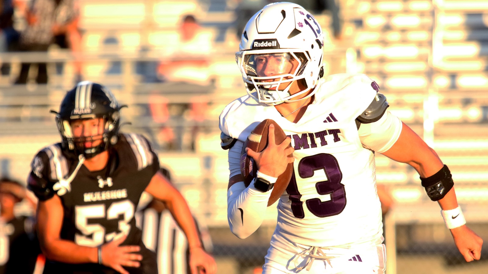 MIT football player in white jersey number 13 runs with the ball during a game on a sunny day, pursued by a defender in black.