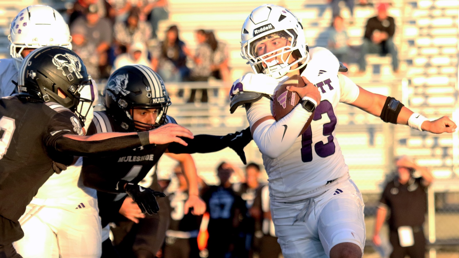 Quarterback in white jersey (number 13) runs with the ball while defenders in black reach in on a football field.