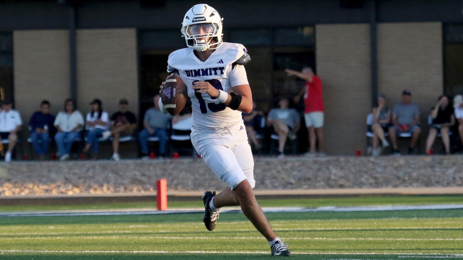 Football player in a white uniform with number 13 runs with the ball during a game, spectators seated in the background.