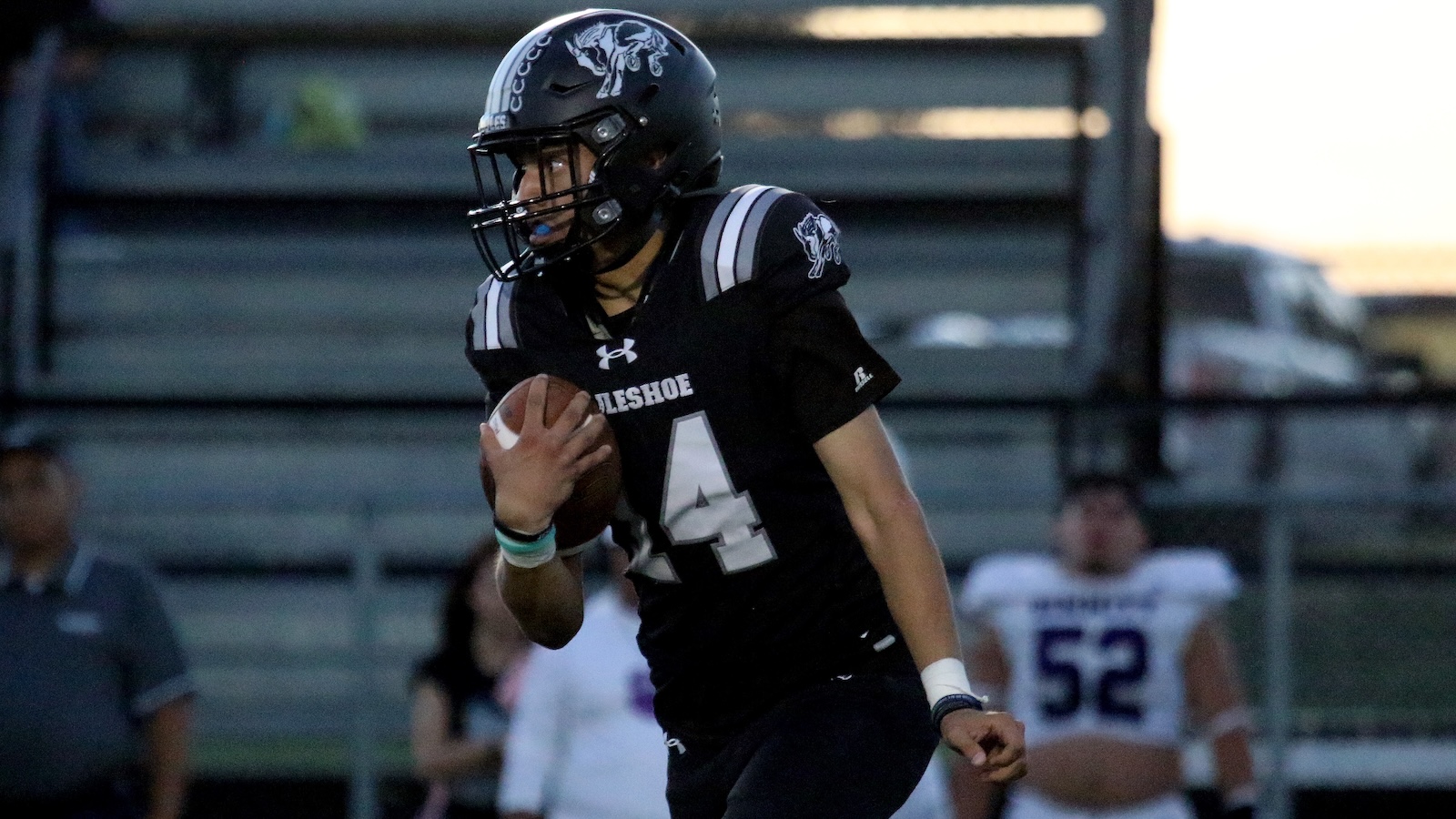 American football player in a black uniform with number 44 runs with the ball during a game, wearing a helmet with a mascot logo and dark facemask, bleachers in the background.