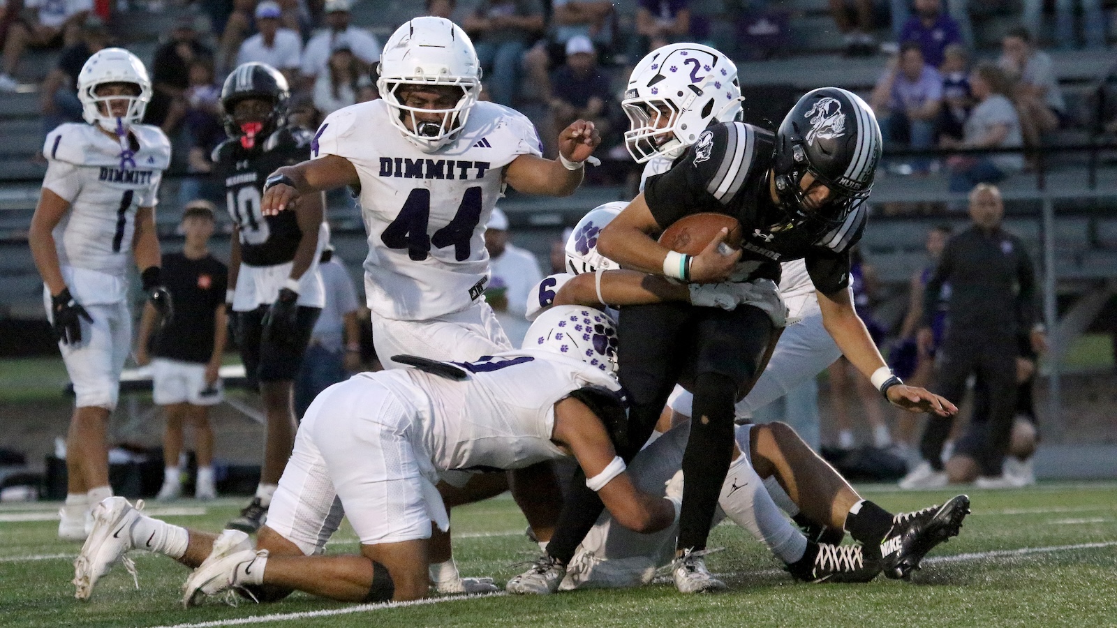 Black-uniformed football player carries ball as he is tackled by white-uniformed defenders on a grassy field during a game with spectators in the stands behind them.