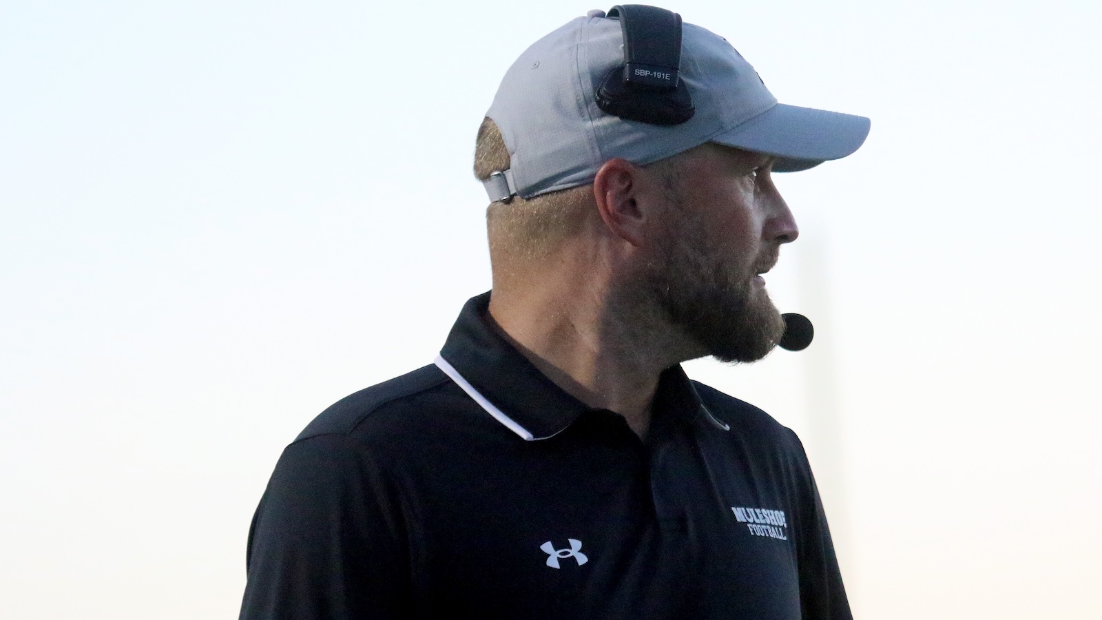 Side profile of a football coach wearing a gray cap and headset microphone, addressing players.