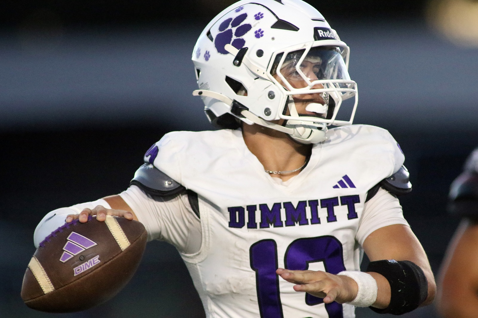 Football player in a white Dimmitt jersey (#10) with a purple number, holding a ball and preparing to pass, wearing a white helmet with purple paw graphic.