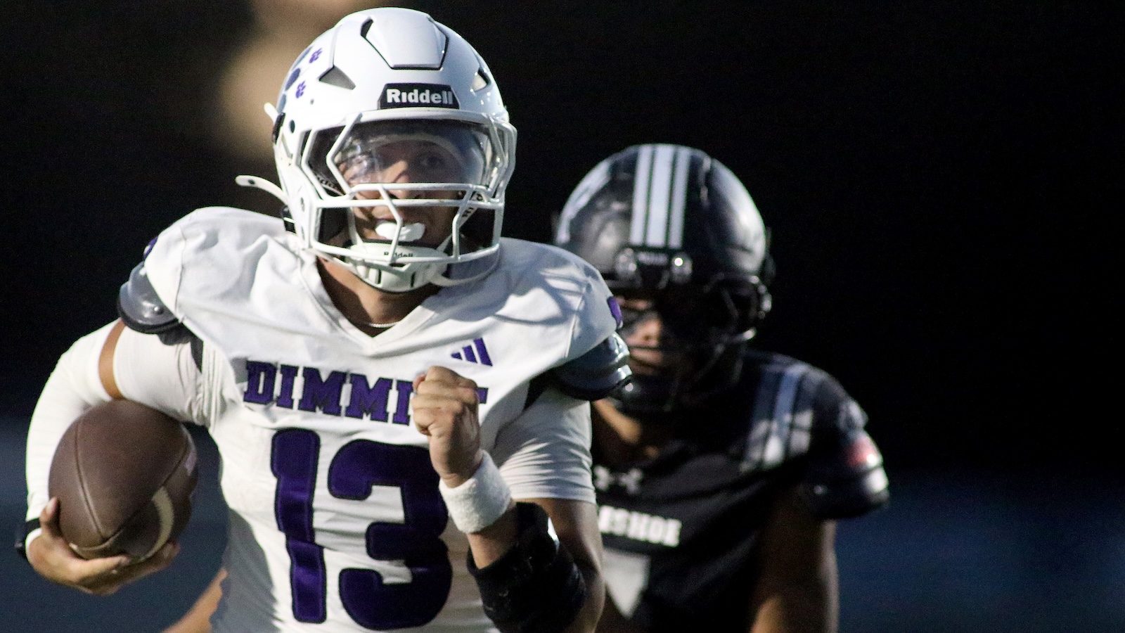 Football player in white uniform (number 13) runs with the ball during a night game, defender in dark jersey pursuing behind him