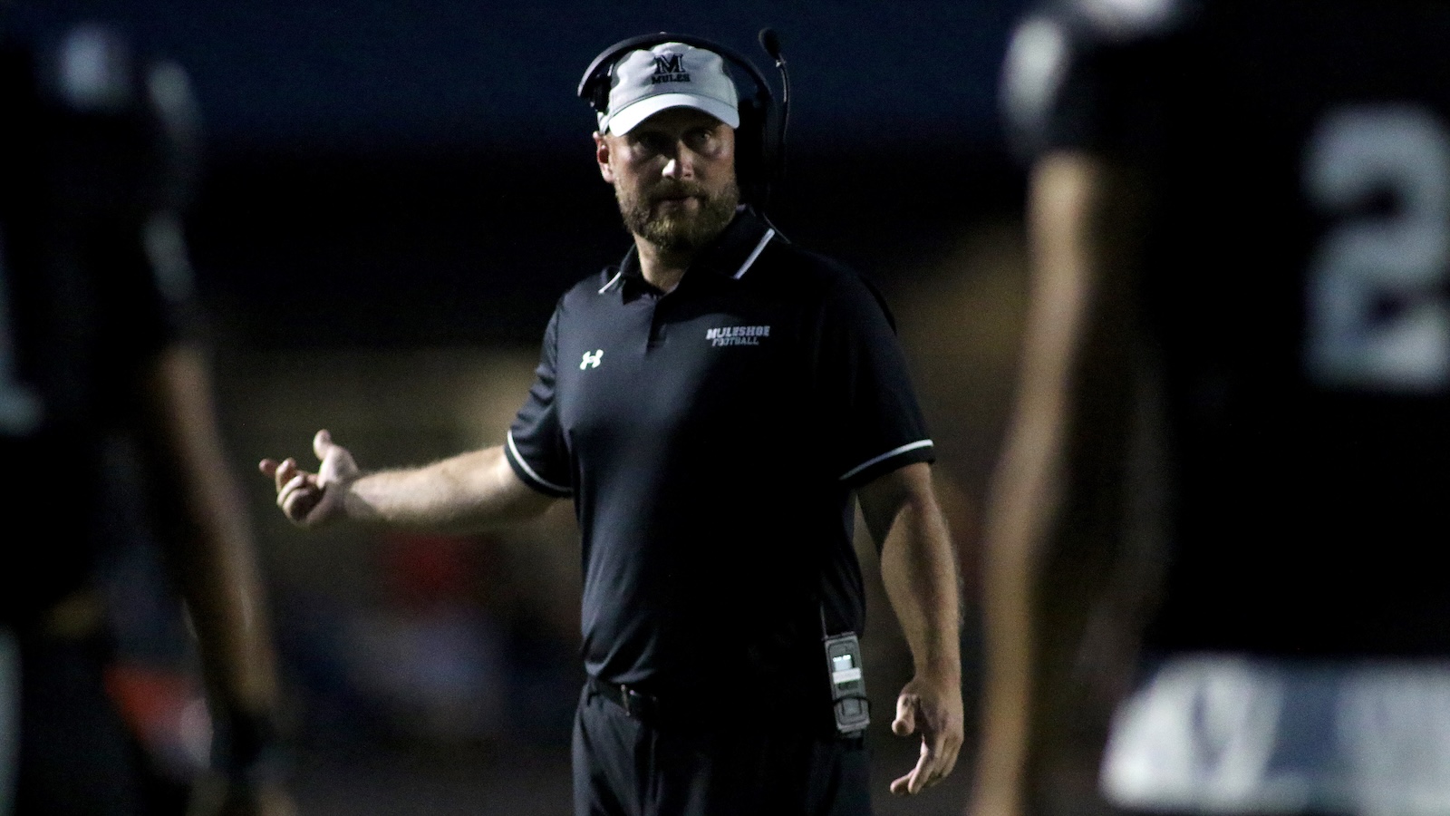 Football coach in a black polo with a headset, standing on the sideline and gesturing during a night game.