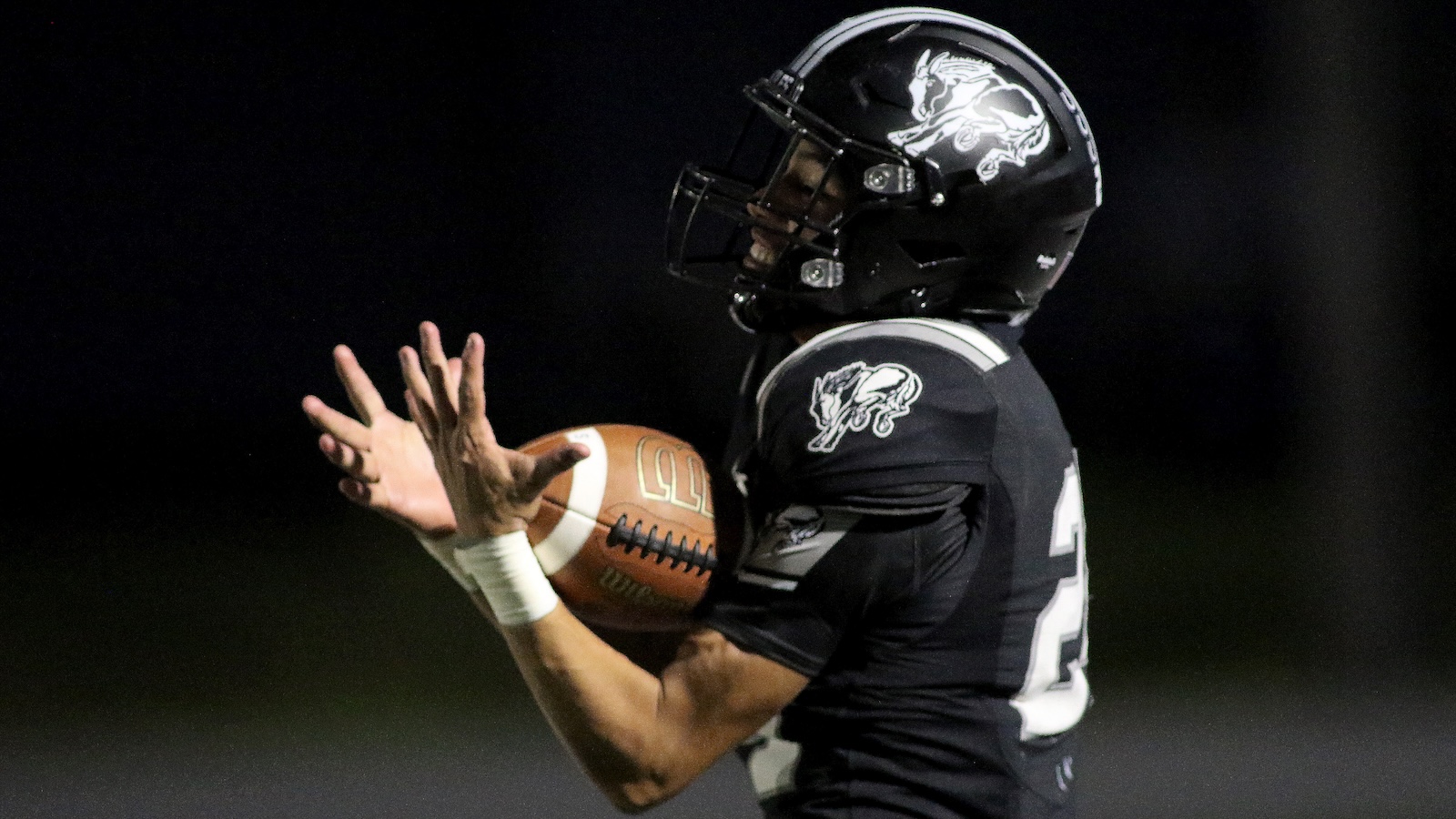 Football player in a black jersey reaches to catch a football at night under stadium lights, with a focused pose.