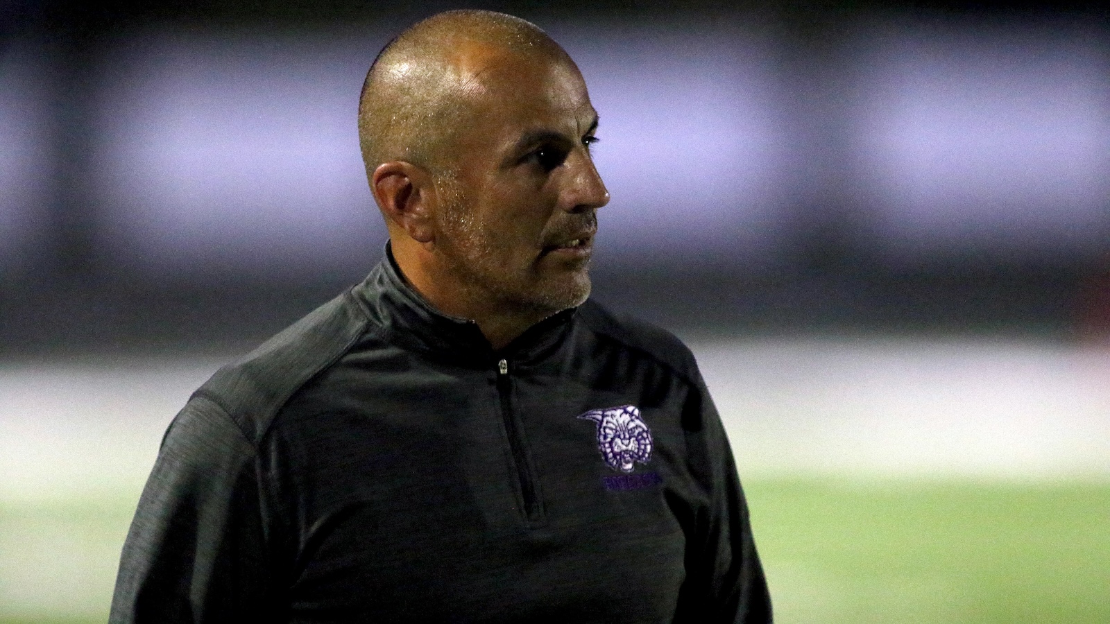 Man with a shaved head in a black athletic jacket looking to the right on a field.