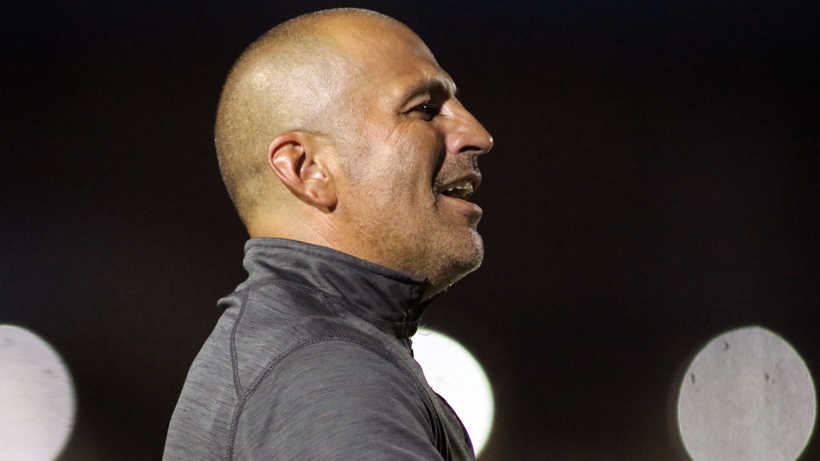 Profile of a bald man in a gray athletic jacket, facing right, against a dark background with blurred lights.