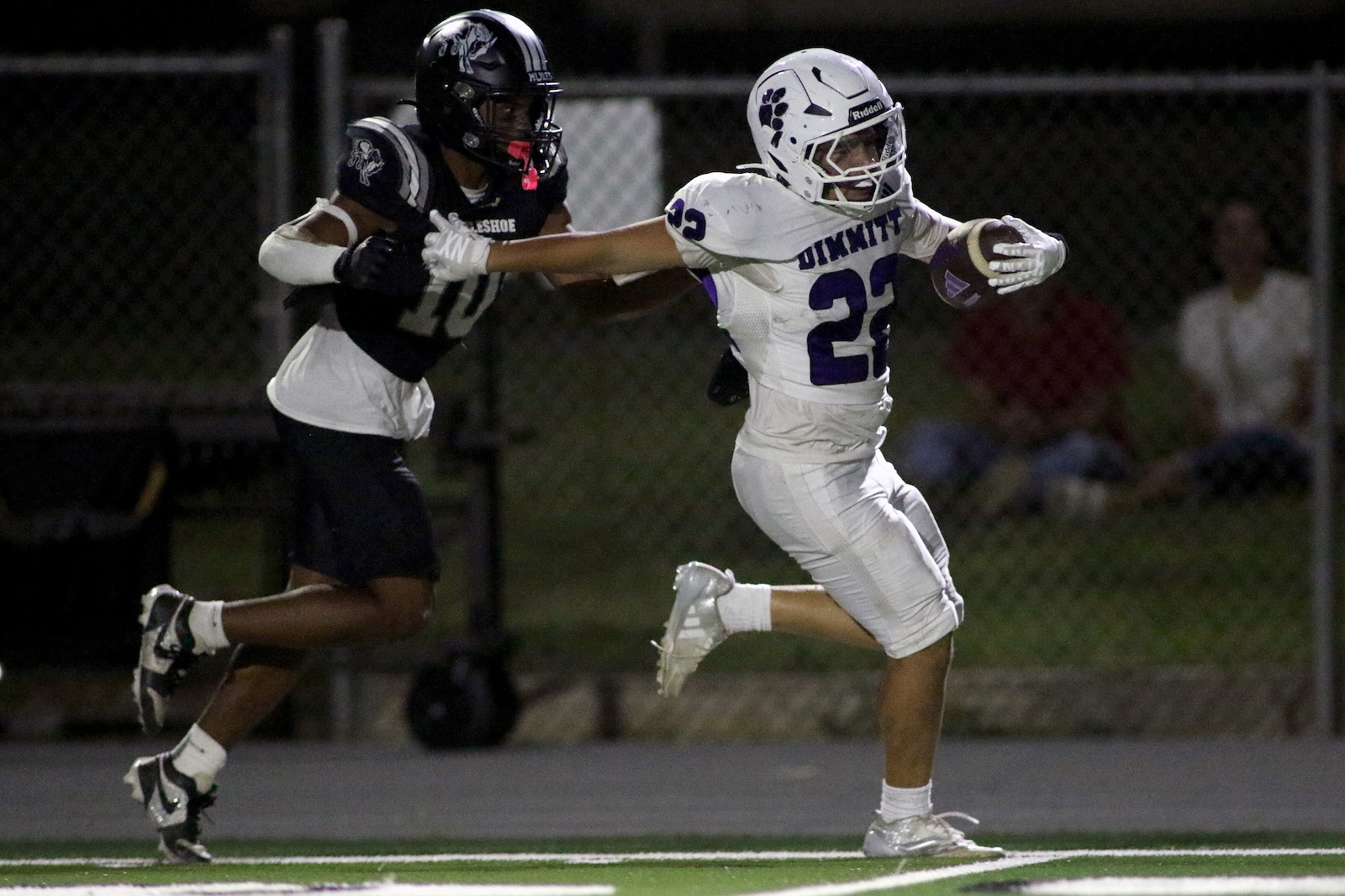 Athlete in white uniform with number 23 runs with ball while a tackler in black jersey pursues at night on a football field