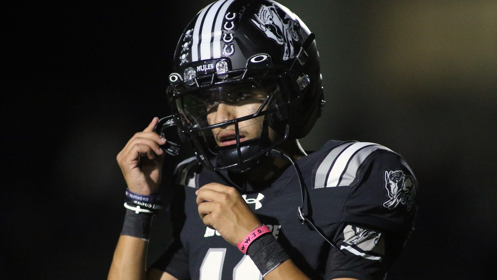 Football player in a dark uniform adjusts his facemask during a game, wearing a black helmet with white stripes and a logo on the sleeve.